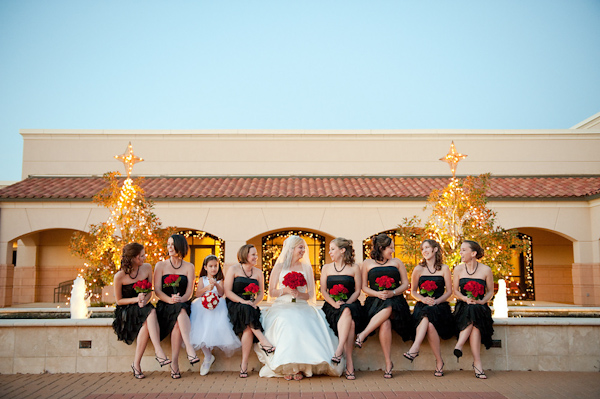 the bride in white with her bridesmaids in black and flower girl in white sitting on the edge of a fountain holding red rose bouquets - on the photo by Houston based wedding photographer Adam Nyholt 
